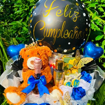 Cesta de regalo con osito, globo de cumpleaños, botella y detalles en azul y amarillo.