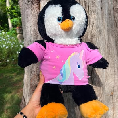 Peluche de pingüino con camiseta rosa y dibujo de unicornio.