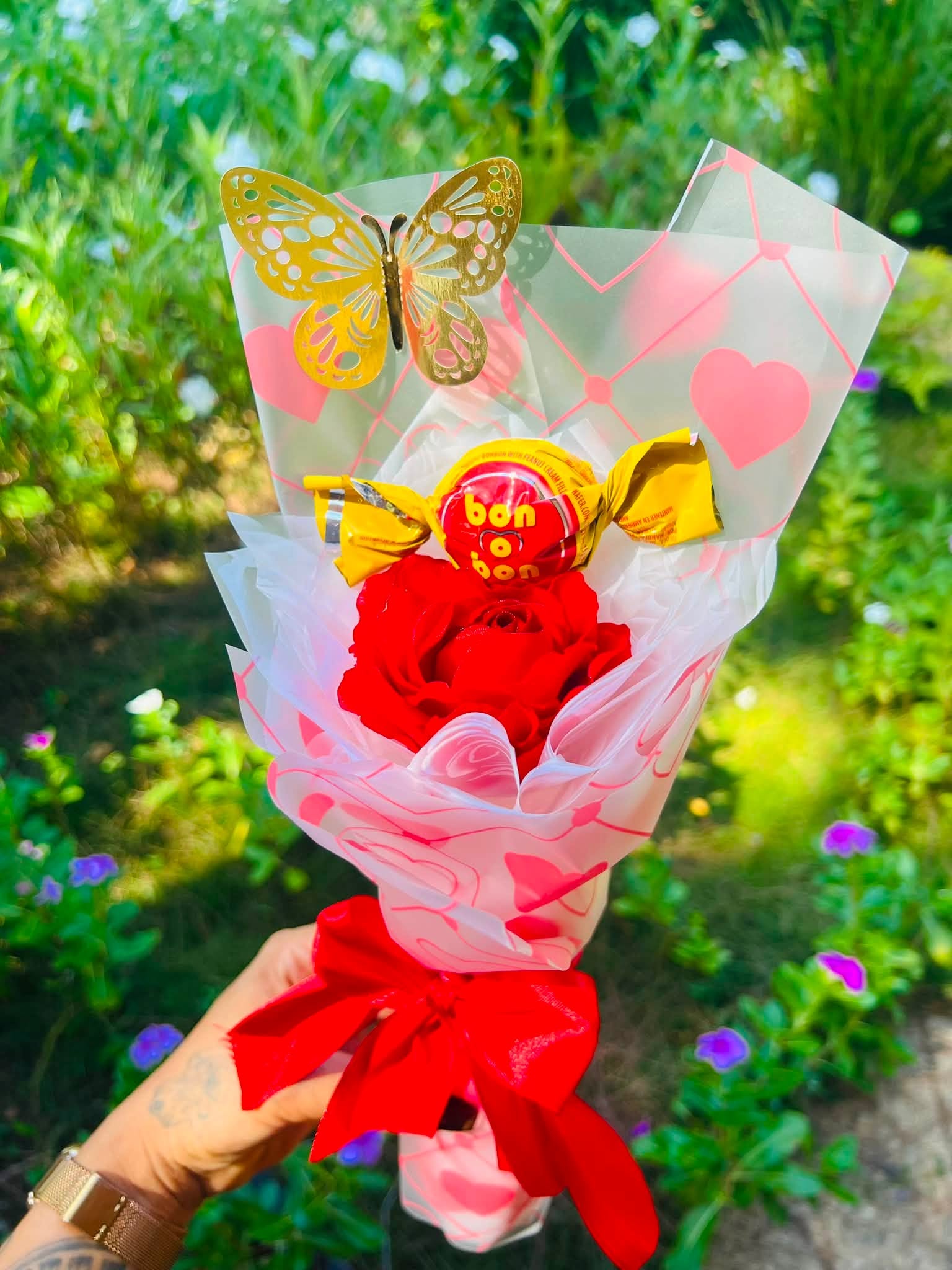 Rosa roja, bombón y mariposa dorada en envoltura de corazones.