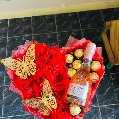 Arreglo de rosas rojas, bombones, mariposas doradas y una botella como complemento.