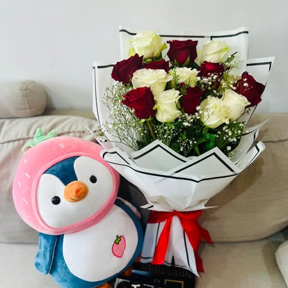 Ramo de rosas rojas y claras junto a un pingüino de peluche con capucha rosa.