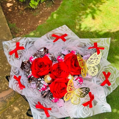 Ramo de rosas rojas con bombones y mariposas, envuelto en papel claro con lazos rojos.